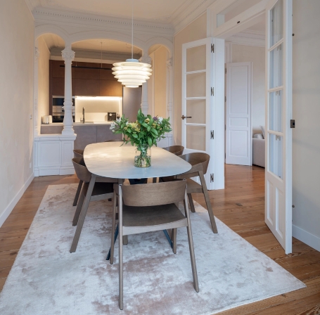 Comedor con mesa blanca, sillas de madera y lámpara de diseño frente a arcos clásicos.