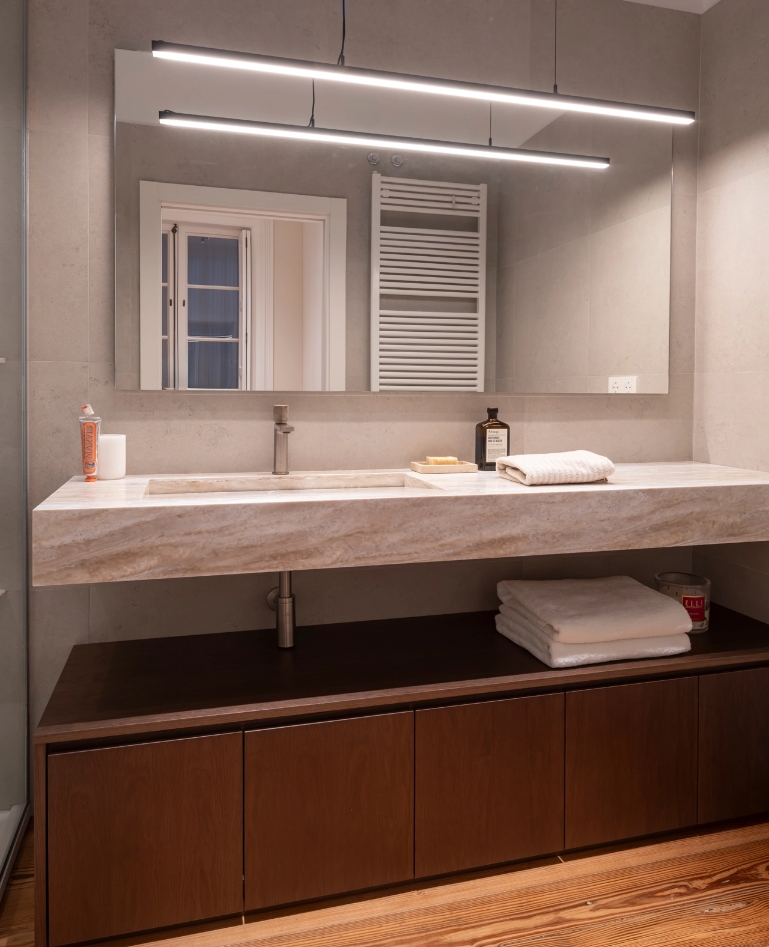 Baño con lavabo de piedra sobre mueble de madera, espejo amplio y luces lineales.