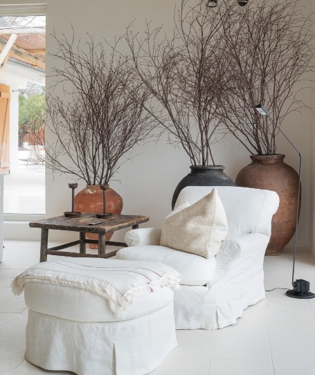 Reading corner with white armchair, footrest, antique side table and large pots with dry branches.
