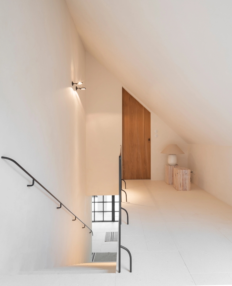 Attic hallway with white walls, wooden door, table lamp and fine metal railing.
