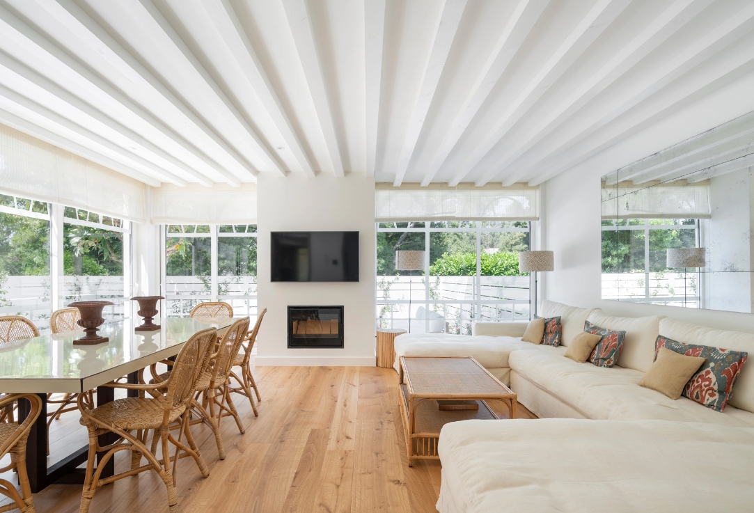 Bright living room with exposed beams, fireplace under TV, white sofa and table with wicker chairs.
