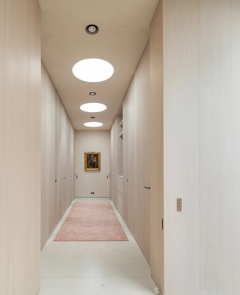Minimalist hallway clad in light wood with circular skylights and elongated pink carpet.