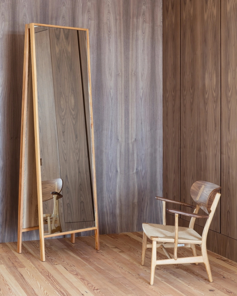 Standing mirror with wooden frame next to design chair and paneled wall.