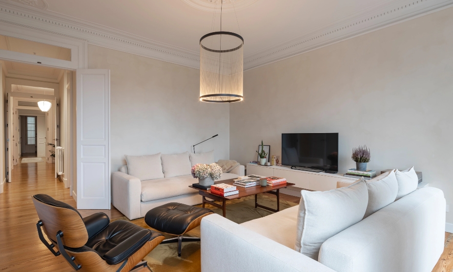 Salón moderno con sofás blancos, mesa de centro de madera y lámpara cilíndrica.