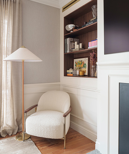 Rincón acogedor con un sillón de tela clara, una lámpara de pie y estanterías llenas de libros.