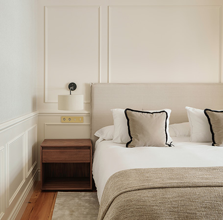 Dormitorio con cama de cabecero claro, mesita de noche de madera y una lámpara de pared.