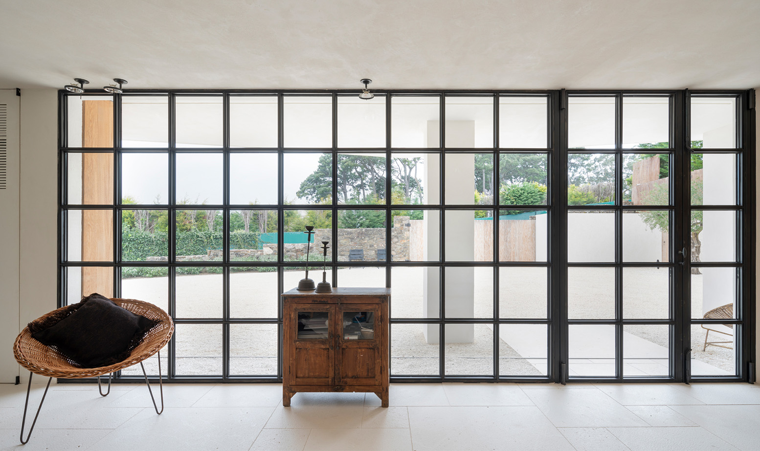 Amplio ventanal industrial de cuadrícula con vista exterior, sillón de mimbre y mueble bajo de madera rústica.
