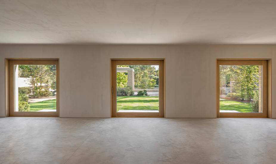 Un espacio interior minimalista con suelo de hormigón pulido y tres grandes puertas de cristal enmarcadas en madera.
