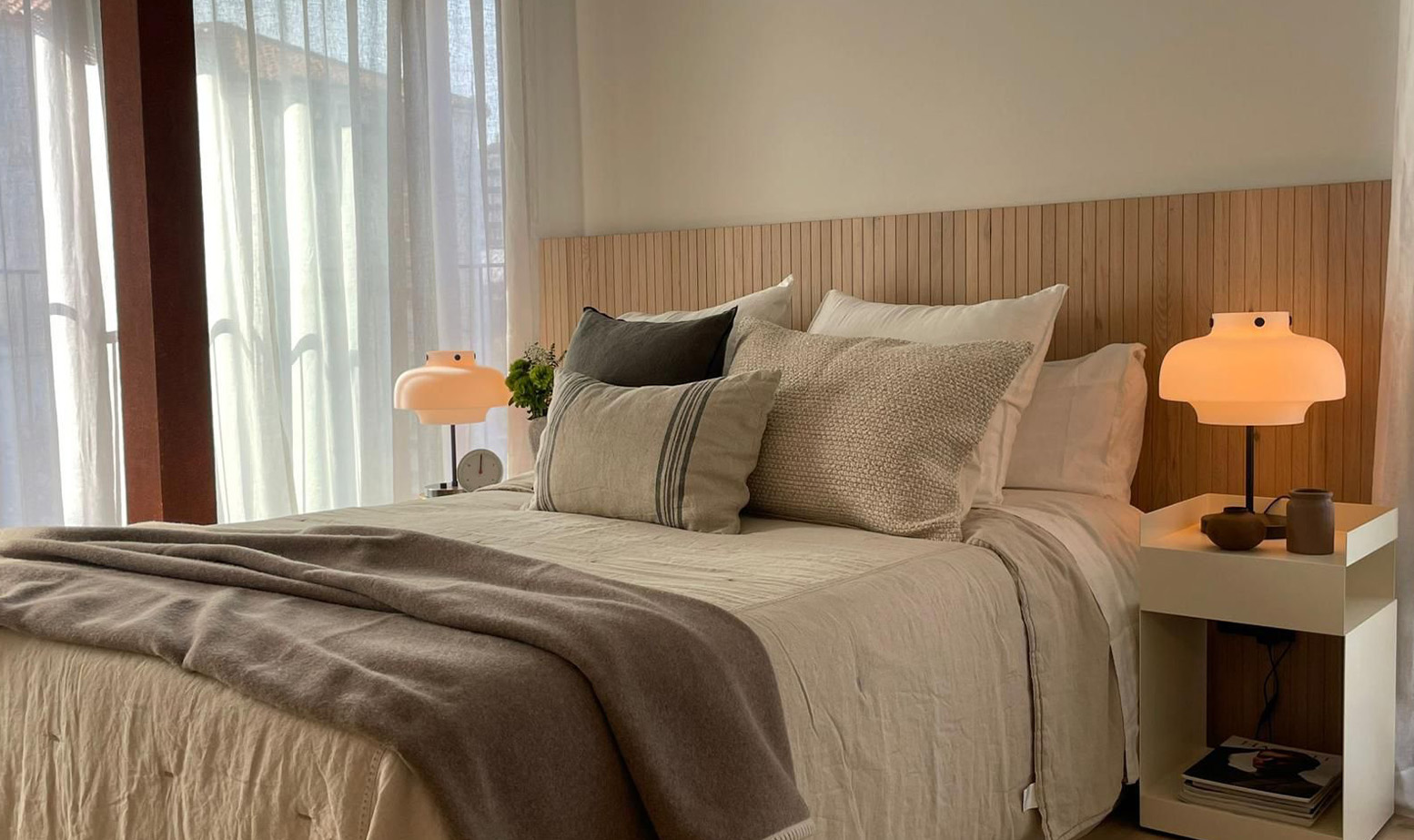 Dormitorio acogedor con cabecero listonado, lámparas cálidas, textiles neutros, cojines decorativos, cortinas ligeras y atmósfera relajante contemporánea.