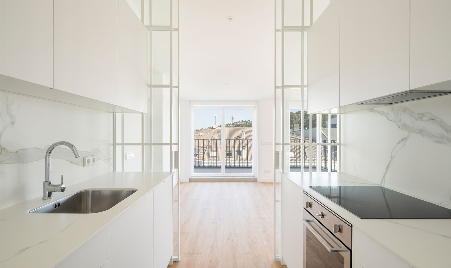 Cocina moderna y blanca vista en perspectiva hacia un salón luminoso, separada por elegantes paneles acristalados de marco blanco.