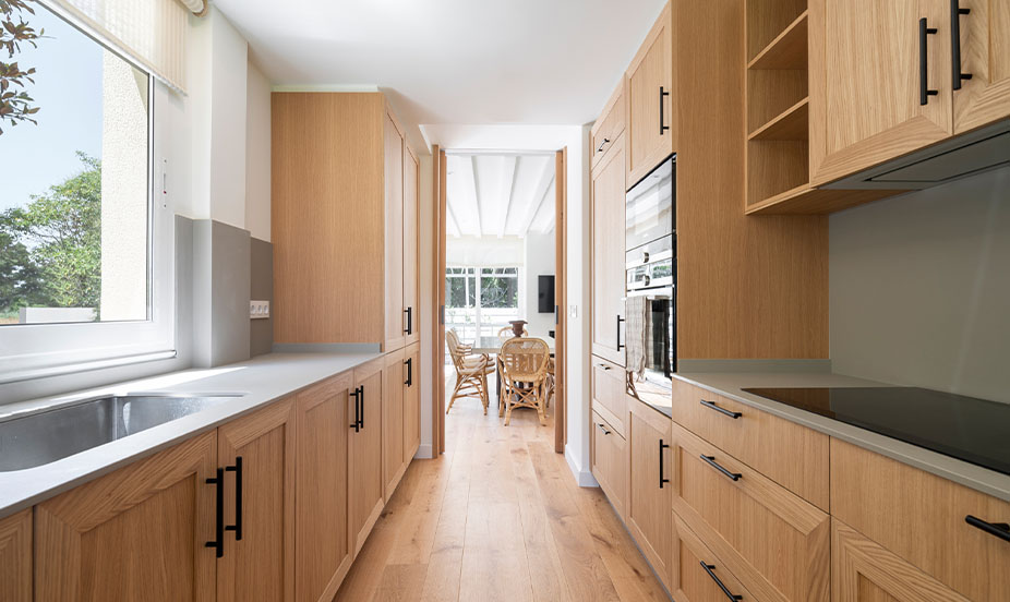 Cocina alargada con muebles de madera hasta el techo, encimera gris, ventana luminosa y pasillo hacia un salón al fondo.