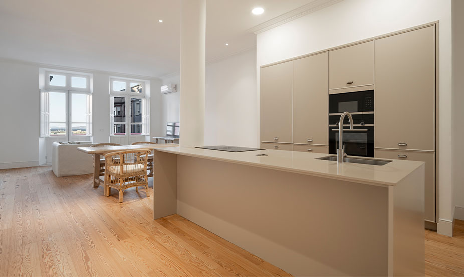 Espacio abierto con suelo continuo de madera, península beige de cocina, zona de comedor y grandes ventanales al fondo