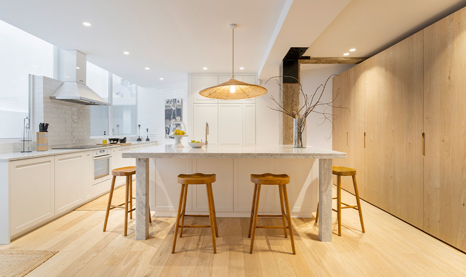 Cocina amplia y luminosa con gran isla de mármol, taburetes de madera, suelo de parquet y armarios integrados al fondo.