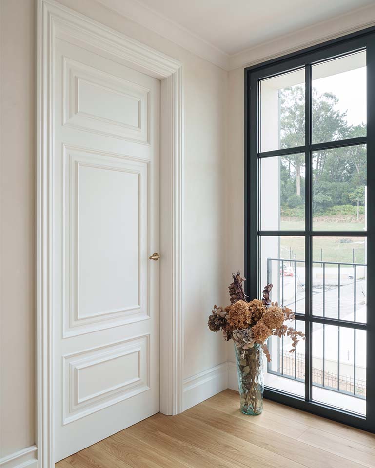 A white-lacquered wooden interior door with classic moldings and gold-colored hardware next to a large window.